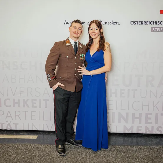 Ein Mann in Militäruniform und eine Frau in einem blauen Kleid stehen eng beieinander vor einer weißen Wand mit Text.
