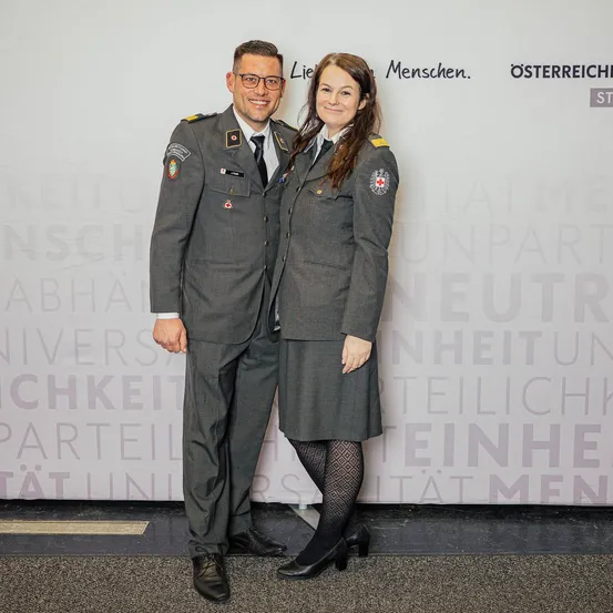 Ein Mann und eine Frau in Militäruniformen stehen eng nebeneinander vor einer großen Wand mit politischem Text im Hintergrund.