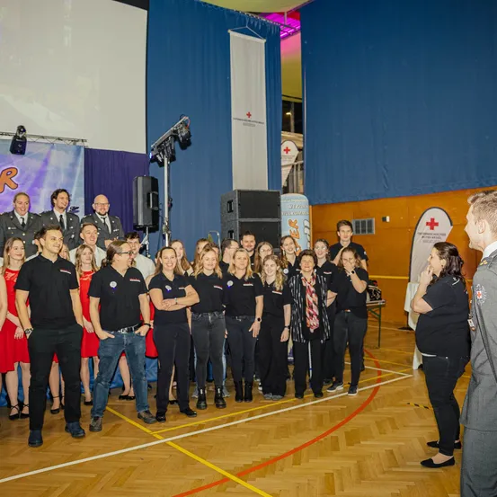Eine Gruppe von Menschen steht in einer Turnhalle. Sie tragen Uniformen und sind in Reihen organisiert. Einige haben Abzeichen auf ihren Hemden. Hinter ihnen stehen Lautsprecher und ein Banner.