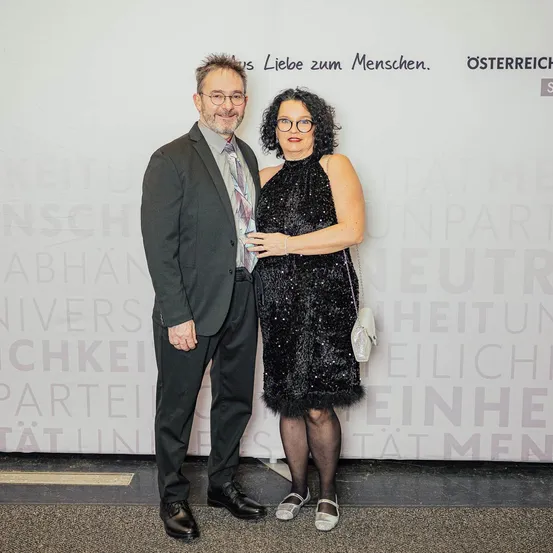 Ein Mann und eine Frau stehen eng beieinander und lächeln für ein Foto vor einer Wand mit einem Slogan, der 'is Liebe zum Menschen' lautet.