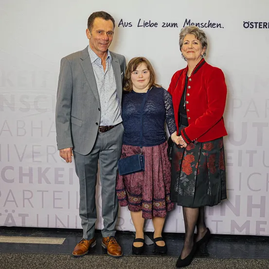 Drei Personen, zwei Frauen und ein Mann, stehen eng beieinander vor einem Banner mit Text. Der Mann trägt einen Anzug und Lederschuhe. Die Frauen tragen Kleider und eine hat eine Handtasche.