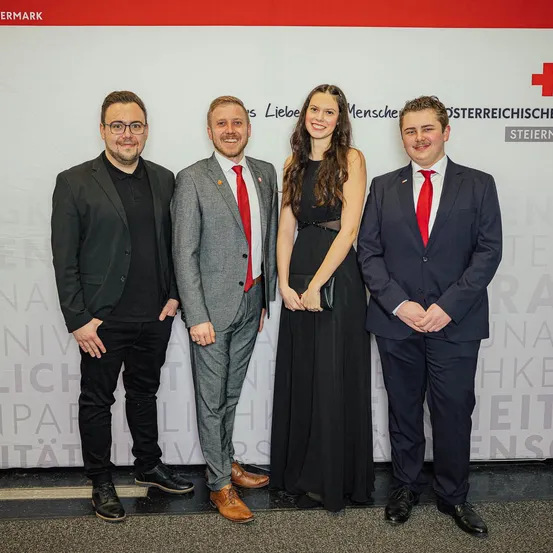 Vier Personen in formeller Kleidung stehen vor einem weißen Hintergrund mit rotem Kreuz und Text, vermutlich bei einer offiziellen Veranstaltung.