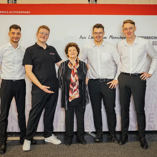 Fünf Personen stehen in einer Reihe vor einem weißen Banner mit rotem Text, in formeller Kleidung. Die Frau in der Mitte ist älter und trägt eine Krawatte. Die beiden Männer an den Seiten tragen Brillen.