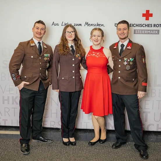 Vier Personen in formeller Kleidung, darunter drei in Militäruniformen, stehen vor einer weißen Wand mit rotem Text und Logos.