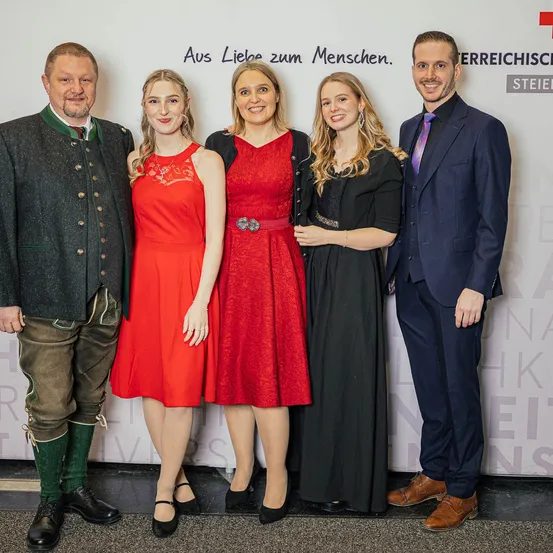 Fünf Personen, feierlich gekleidet, stehen zusammen vor einer Wand. Drei Frauen und zwei Männer, lächelnd. Die Frau links trägt ein rotes Kleid. Die Wand zeigt weißen Text.