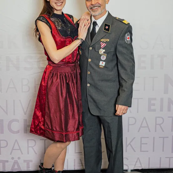Ein Mann in Uniform steht neben einer Frau in einem roten Kleid. Sie scheinen beide für ein Foto zu lächeln.