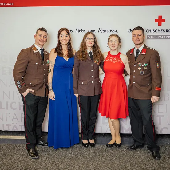 Fünf Personen stehen vor einem Hintergrund mit deutschem Text, lächeln und posieren für ein Foto. Vier tragen braune Uniformen, eine ist in einem roten Kleid.