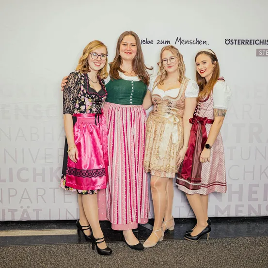 Vier Frauen in traditioneller deutscher Kleidung posieren für ein Foto vor einer weißen Wand mit Text.