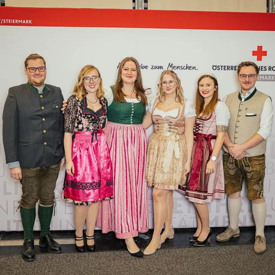 Eine Gruppe von Menschen in traditioneller deutscher Kleidung, die vor einem Banner mit einem roten Kreuz für ein Foto posieren.