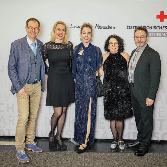 Fünf Menschen stehen lächelnd zusammen für ein Foto. Ein Banner hinter ihnen hat ein rotes Kreuz und steht 'Osterreichisches Steiermark'.