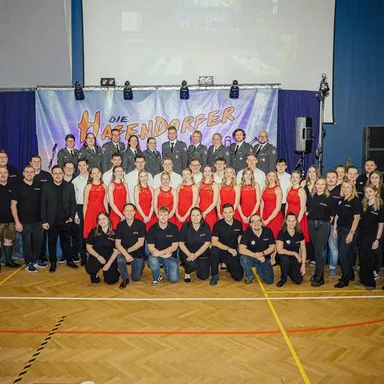 Eine Gruppe von Menschen in Uniform und Freizeitkleidung posiert für ein Foto in einer Halle mit einem Banner, das Die Haenddorfer liest.