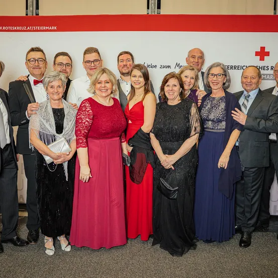Eine Gruppe formell gekleideter Menschen steht zusammen und lächelt für ein Foto. Sie stehen vor einer Wand mit einem roten Kreuz und einer Website-Adresse.