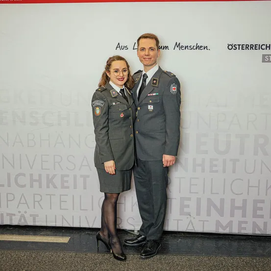 Ein Mann und eine Frau in Militäruniformen stehen vor einem weißen Hintergrund mit österreichischen Texten und Logos. Sie lächeln beide für ein Foto.