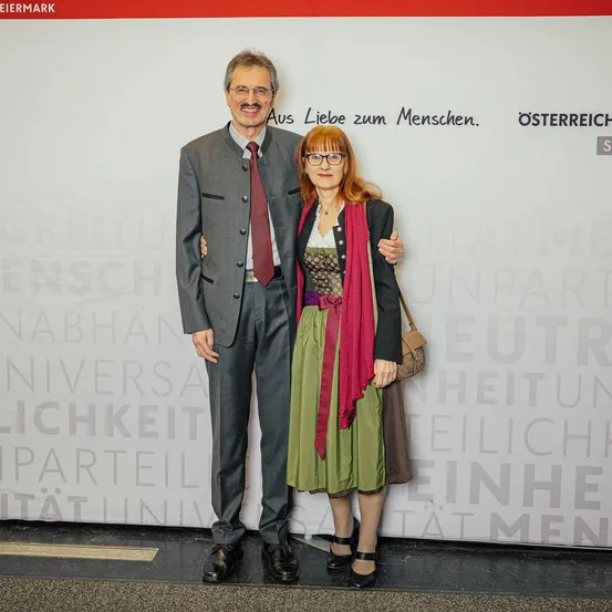 Ein Mann und eine Frau stehen eng nebeneinander vor einer weißen Wand mit deutschem Text. Der Mann trägt einen Anzug und eine Krawatte, während die Frau ein Kleid und eine Brille trägt. Sie lächeln beide.