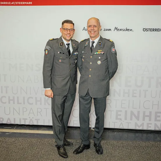 Zwei Männer in Militäruniformen stehen nebeneinander und lächeln für ein Foto vor einem weißen Hintergrund mit verschwommenem Text.