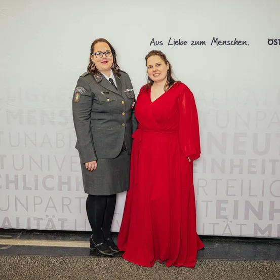 Zwei Frauen stehen nebeneinander, eine in einem roten Kleid und die andere in einer Militäruniform, lächeln für ein Foto vor einem weißen Banner mit deutschem Text.