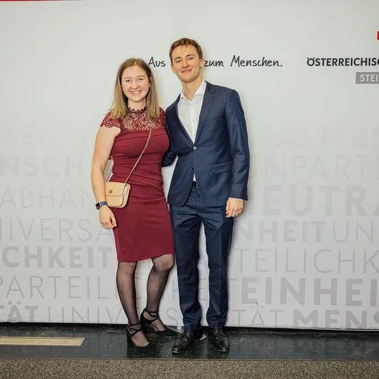 Ein Mann und eine Frau stehen lächelnd eng beieinander vor einer Wand mit Text in einer fremden Sprache. Der Mann ist in einem dunklen Anzug und die Frau in einem roten Kleid.