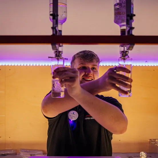 Ein lächelnder Barkeeper hält zwei mit Eis und Flüssigkeit gefüllte Gläser, mit Flaschen darüber, unter einer gelben Wand mit lila Lichtern.