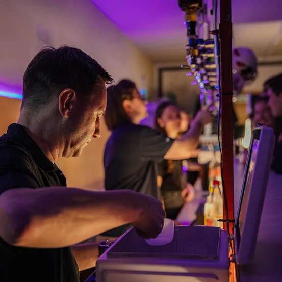 Ein Mann in einem schwarzen Shirt arbeitet hinter einer Bartheke, umgeben von Flaschen und einem Helm. Eine Frau hinter ihm ist beschäftigt, während andere in der Nähe plaudern. Die Bar ist mit lila Lichtern schwach beleuchtet.