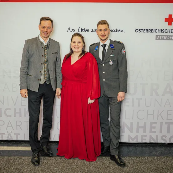 Drei Personen stehen vor einem weißen Hintergrund mit deutschem Text. Der Mann links trägt einen grauen Anzug, die Frau in der Mitte ein rotes Kleid und der Mann rechts eine Militäruniform.