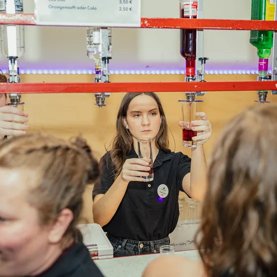 Eine Frau in einem schwarzen Shirt hält ein Glas mit roter Flüssigkeit vor andere. Über ihr sind Flaschen mit Getränkeoptionen beschriftet.