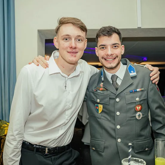 Zwei Männer stehen eng beieinander und lächeln für ein Foto. Einer in einem weißen Hemd, der andere in einer Militäruniform mit Medaillen. Sie sind in einem Raum mit einem blauen Vorhang und einem Glas mit einem Strohhalm.