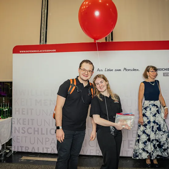 Drei Personen stehen vor einem großen weißen Banner mit Text. Ein Mann und eine Frau halten einen großen roten Ballon. Eine Frau in einem Kleid hält ein kleines Paket. Hinter ihnen ist ein Tisch mit grünen Flaschen zu sehen.