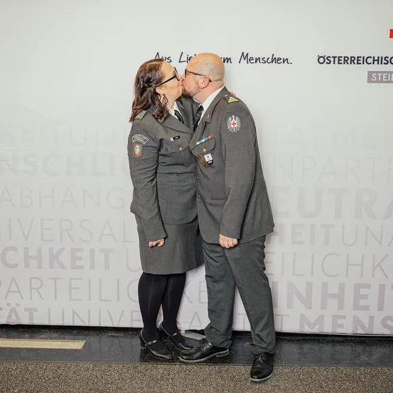Ein Mann und eine Frau in Militäruniformen stehen nah beieinander und küssen sich. Sie stehen vor einer weißen Wand mit blauem Text.