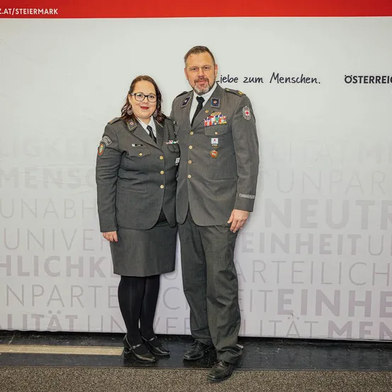 Ein Mann und eine Frau in Militäruniformen stehen nebeneinander vor einer Wand mit Text.