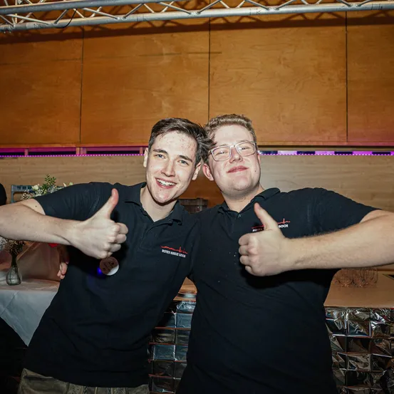 Zwei Männer stehen nebeneinander in einer Partyumgebung, beide tragen schwarze Polo-Shirts mit Logos und zeigen Daumen hoch. Hinter ihnen befindet sich eine Holzwand mit lila Beleuchtung.