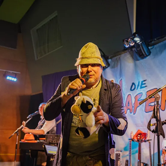 Ein Mann auf der Bühne hält ein Mikrofon und ein Plüschtier. Hinter ihm spielt ein Musiker an einem Keyboard. Ein Banner mit dem Wort 'Die' ist sichtbar.