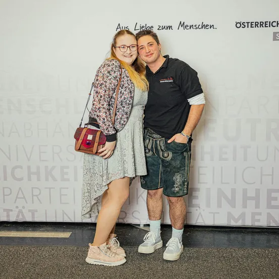 Ein lächelndes Paar steht gemeinsam vor einer weißen Wand mit großen Buchstaben. Die Frau trägt eine Brille, eine Blumenjacke und Turnschuhe. Der Mann trägt ein schwarzes T-Shirt, grüne Shorts und weiße Socken.