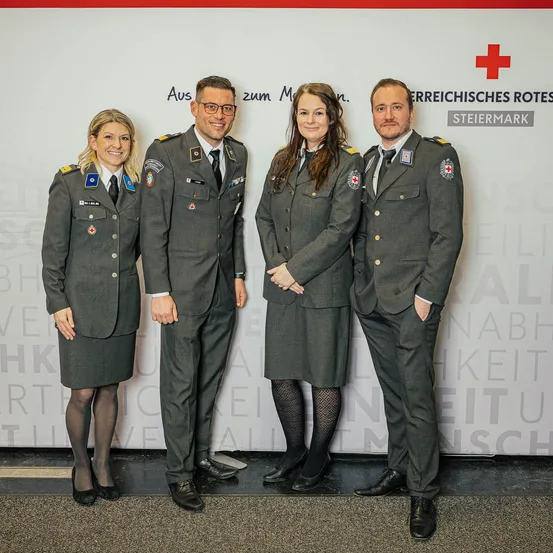 Vier Personen in Uniform stehen vor einer weißen Wand mit rotem Text und lächeln für ein Foto. Darunter sind zwei Frauen und zwei Männer.