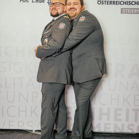 Zwei Männer in Militäruniformen posieren für ein Foto vor einem weißen Hintergrund mit deutschem Text. Einer trägt eine Brille, und beide haben lächelnde Gesichter.