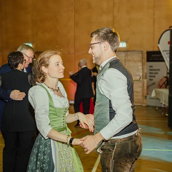 Ein Mann und eine Frau in traditioneller bayerischer Kleidung tanzen in einer Halle, während andere zusehen. Dahinter beobachtet eine Person in formeller Kleidung.