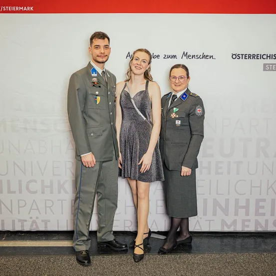 Drei Menschen stehen vor einer Wand mit Text, in Uniformen gekleidet. Ein Mann links, eine Frau in einem Kleid in der Mitte und eine Frau in einer Uniform rechts.