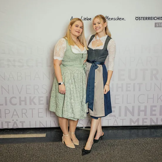 Zwei Frauen stehen vor einer Wand mit Text. Beide tragen traditionelle Kleidung, eine in Grün und eine in Blau. Sie scheinen zu lächeln und posieren für ein Foto.