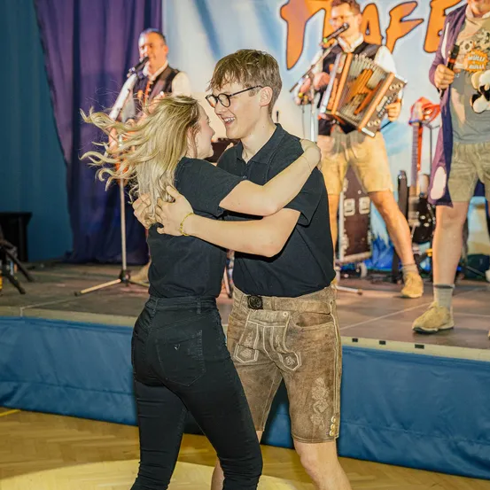 Ein Mann und eine Frau tanzen auf einer Bühne, während eine Band im Hintergrund spielt. Die Band spielt verschiedene Instrumente, und das Publikum schaut ihnen zu.