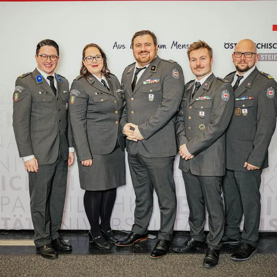 Fünf Personen in gleichen Uniformen stehen in einer Reihe und lächeln für ein Foto. Der Hintergrund zeigt weißen Text auf rotem und weißem Hintergrund.