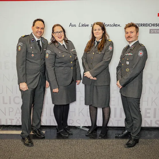 Vier Personen in grauen Uniformen mit verschiedenen Abzeichen und Orden stehen gemeinsam vor einem weißen Hintergrund.