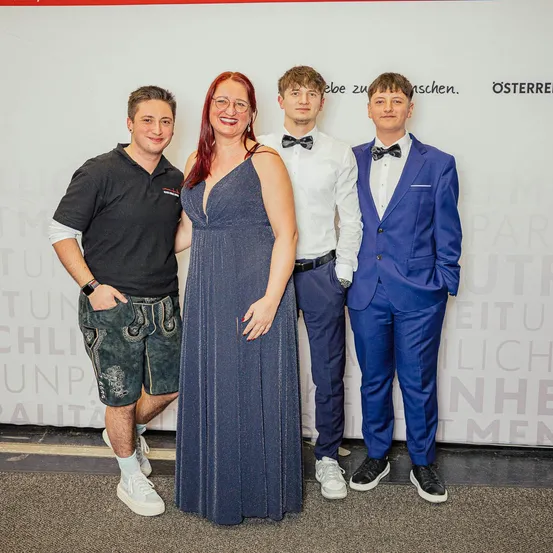 Vier Menschen stehen gemeinsam und lächeln für ein Foto. Die Frau trägt eine Brille und ein Kleid, während die Männer Anzüge und Krawatten tragen. Sie stehen vor einer weißen Wand mit Text.
