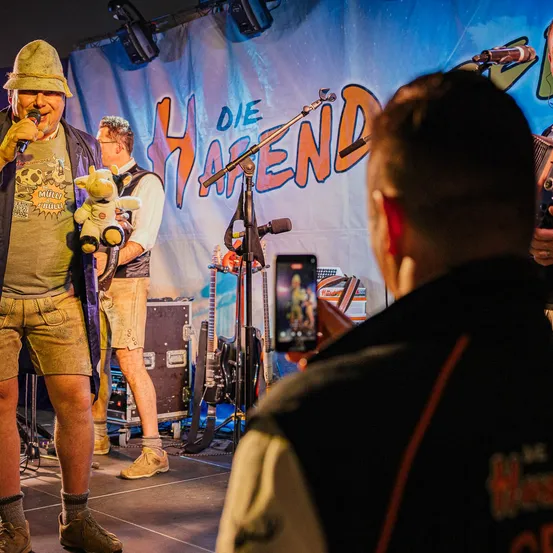 Ein Performer auf der Bühne mit einem Mikrofon hält ein Kuscheltier, während eine Person dahinter ein Foto mit dem Handy macht. Ein Banner mit orangefarbener Schrift steht für 'DIE HAFEND'.