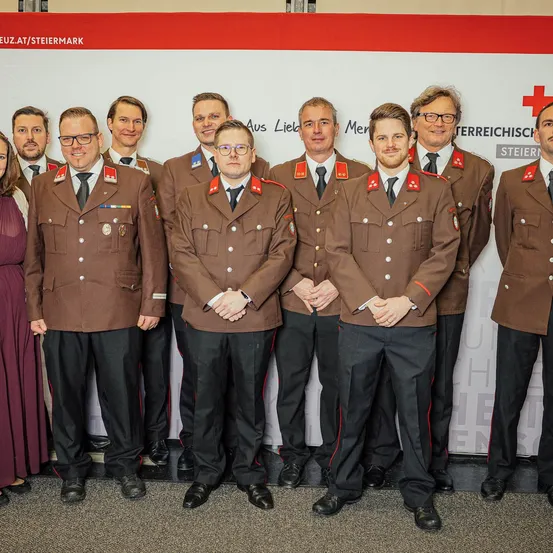 Eine Gruppe von Männern in Militäruniformen steht vor einem weißen Banner. Sie tragen Medaillen und schwarze Krawatten. Sie schauen nach vorne.