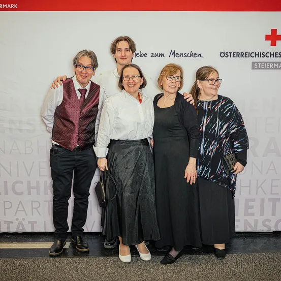 Fünf Personen stehen vor einer weißen Wand mit rotem Rand und lächeln für ein Foto. Sie tragen alle eine Brille. Der Mann links trägt eine Weste und die Frau rechts hält eine Handtasche.