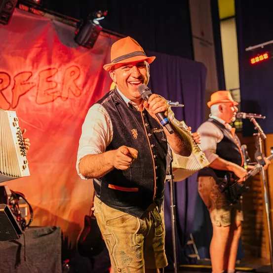 Eine Band tritt auf der Bühne auf, ein Mann singt in ein Mikrofon, während ein anderer die Gitarre spielt. Dahinter ist ein Hintergrund mit rotem Text.