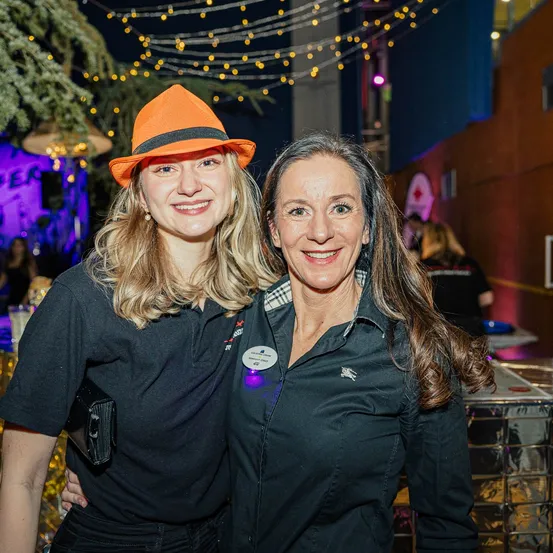 Zwei Frauen in gleichen schwarzen Shirts und Hüten stehen lächelnd für ein Foto. Der Hintergrund zeigt festliche Lichter und eine lila Wand.