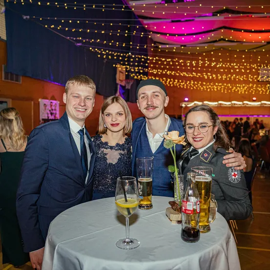 Vier Menschen in formeller Kleidung stehen um einen Tisch mit Getränken. Zwei Männer tragen Anzüge und einer ist in Uniform. Sie lächeln und posieren für ein Foto.