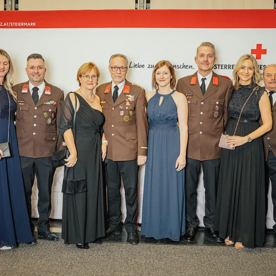 Eine Gruppe von Menschen in formeller Kleidung steht vor einem Hintergrund mit dem Roten Kreuz-Emblem und Text auf Deutsch. Sie scheinen für ein Foto zu posieren.