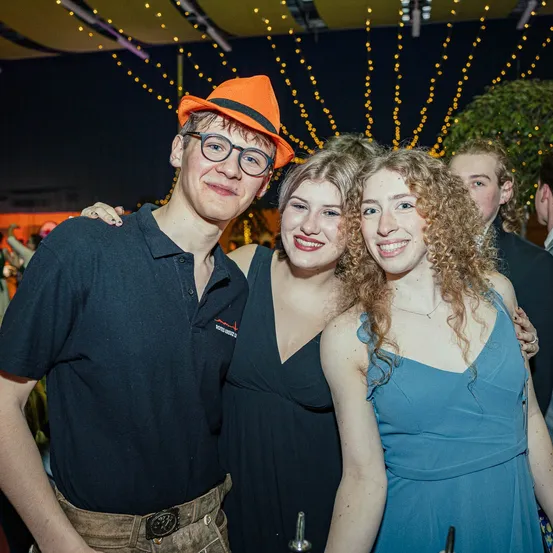 Ein Mann und zwei Frauen posieren für ein Foto auf einer Party, wobei der Mann einen orangefarbenen Hut trägt und die Frauen schwarze und blaue Kleider tragen.