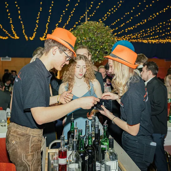 Ein Mann und eine Frau mit orangefarbenen Hüten interagieren miteinander an einer Bar. Sie scheinen sich in einer festlichen Umgebung zu befinden, mit mehreren Leuten im Hintergrund. Flaschen und Gläser stehen auf der Theke.